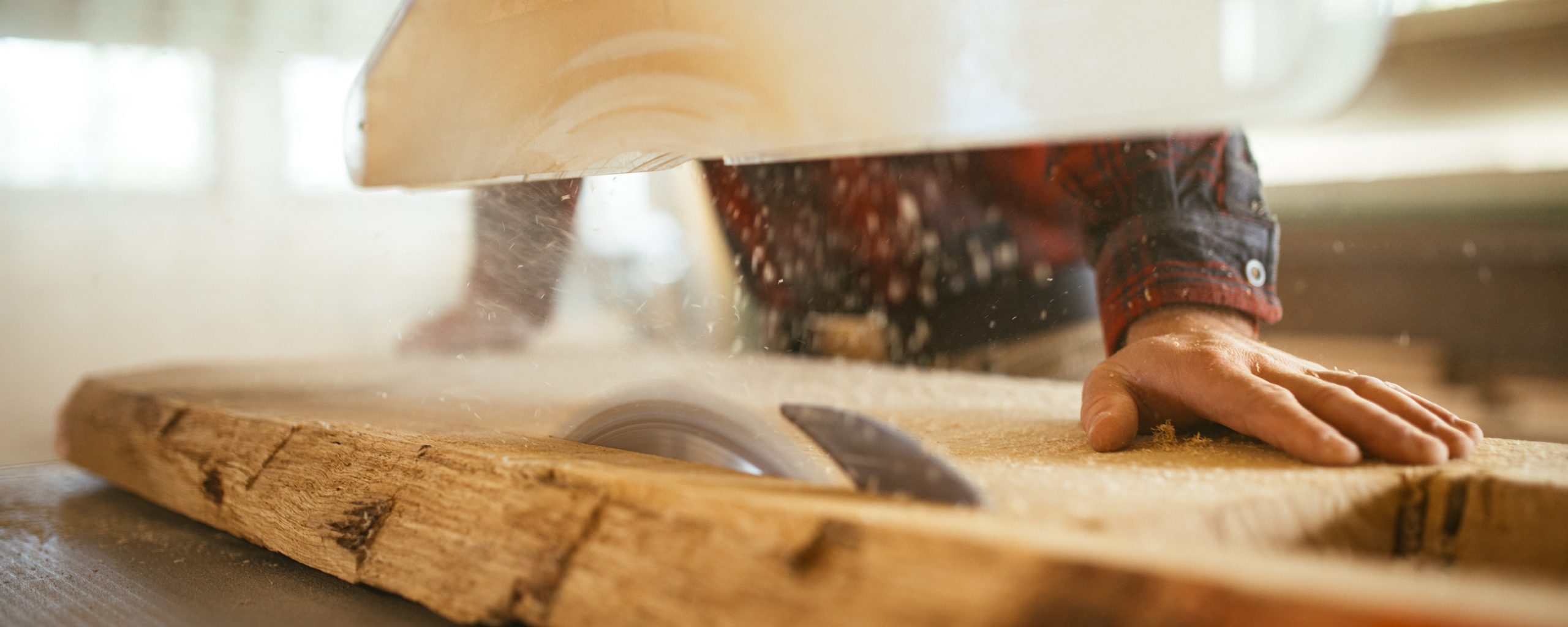 Ein langes, flaches Holzstück wird mit einer Kreissäge bearbeitet. Ein Mann, von dem nur die Hände und ein kariertes Hemd zu sehen sind, hält das Holz fest.